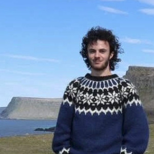 A photo of Sean Brynjólfsson in a beautiful lopapeysa standing
      at Látrabjarg with the westfjords in the background on a clear day.